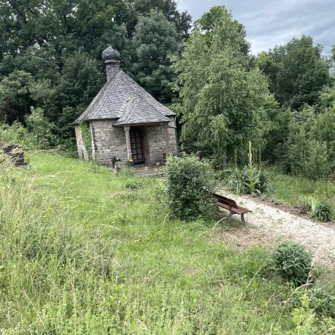 Eine kleine Steinkapelle mit Schieferdach steht inmitten üppiger Vegetation. Ein Kiesweg führt zu einer Holzbank vor der Kapelle., © Daniel Köhler Eine kleine Steinkapelle mit Schieferdach steht inmitten üppiger Vegetation. Ein Kiesweg führt zu einer Holzbank vor der Kapelle., © Daniel Köhler