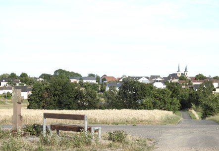 A bench and a cross stand in front of a field. A village with a church and two towers can be seen in the background., &copy; Thomas Neises