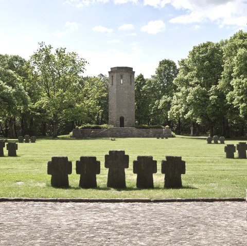 Een oorlogsmonument met stenen kruisen op een weiland, een toren op de achtergrond, omringd door groene bomen onder een blauwe lucht., © TI Bitburger Land