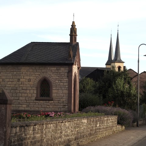 Steinkapelle mit spitzem Dach und buntem Fenster, umgeben von Blumen. Im Hintergrund sind Kirchtürme zu sehen., © Thomas Neises
