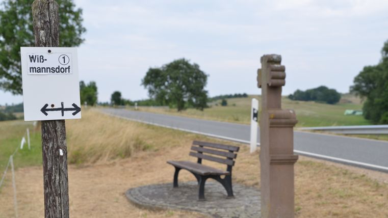 Un panneau indicateur avec l'inscription 'Wißmannsdorf' et un banc à côté d'une route dans un environnement rural.