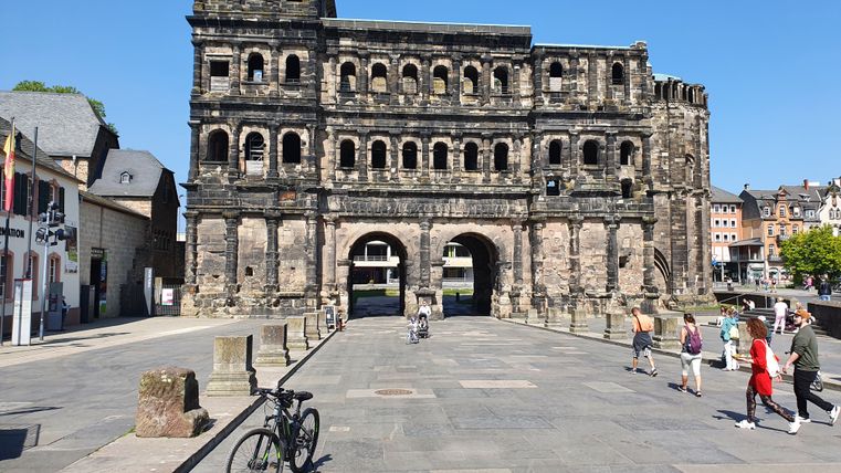 Porta Nigra in Trier, Deutschland, bei sonnigem Wetter mit Passanten und einem Fahrrad im Vordergrund.