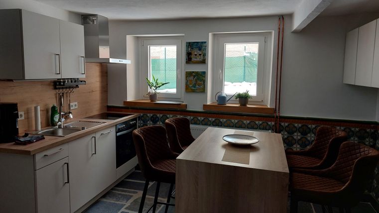 Een moderne keuken met houten elementen en een eettafel. De ramen laten veel licht binnen en er staan groene planten op de vensterbank.