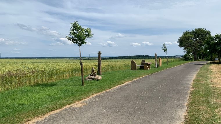 Ein asphaltierter Weg führt durch eine ländliche Landschaft mit Feldern und Bäumen, daneben stehen Bänke und Steinskulpturen.