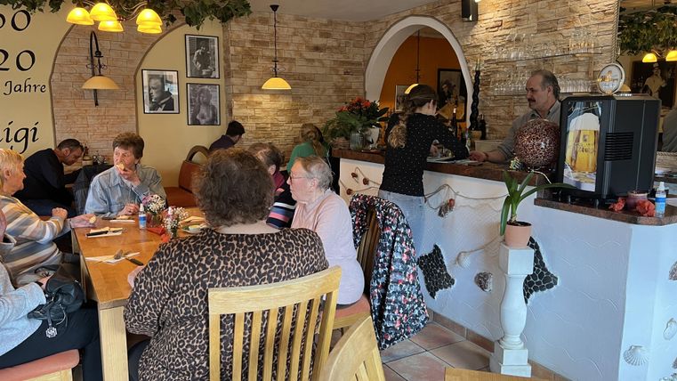 Ein gemütliches Café mit mehreren Gästen, die an Holztischen sitzen. Im Hintergrund bedient eine Person an der Theke.