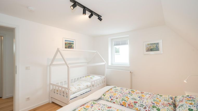 Een lichte slaapkamer met een tweepersoonsbed en een kinderbed in de vorm van een huis. De muren zijn licht gehouden en versierd met afbeeldingen.