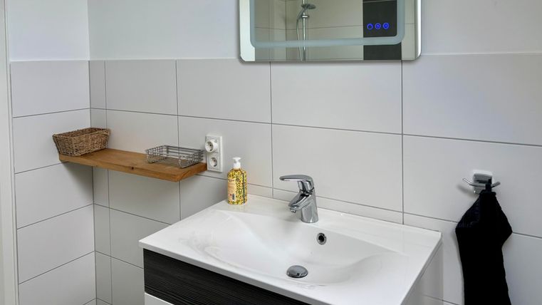 Une salle de bain moderne avec un lavabo, un miroir et des carreaux clairs. Sur une étagère en bois se trouvent du savon et un panier.