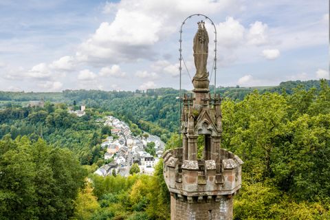 Une statue sur une haute tour surplombe un paysage verdoyant et un petit village dans la vallée. Le ciel est partiellement nuageux et la nature est luxuriante.
