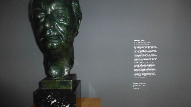 Eine grüne Büste eines Kopfes steht auf einem schwarzen Sockel. Der Hintergrund ist dunkelgrau, und neben der Skulptur befindet sich ein beschriftetes Textfeld.