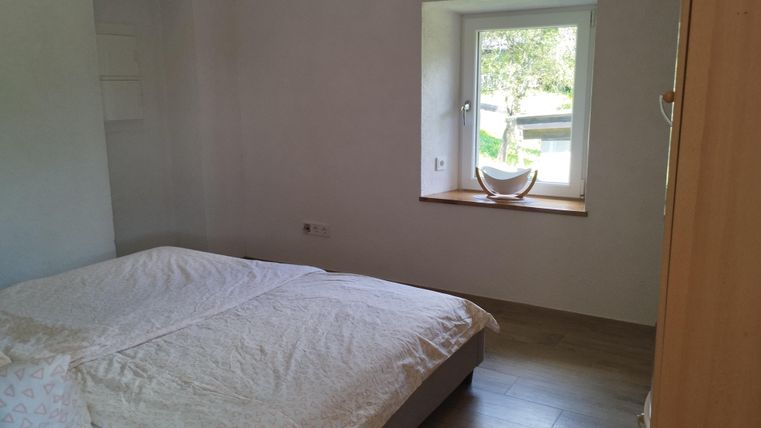 Ein einfaches Schlafzimmer mit einem großen Bett und hellem Tageslicht, das durch ein Fenster strömt. Die Wände sind weiß und der Boden ist aus Holz.