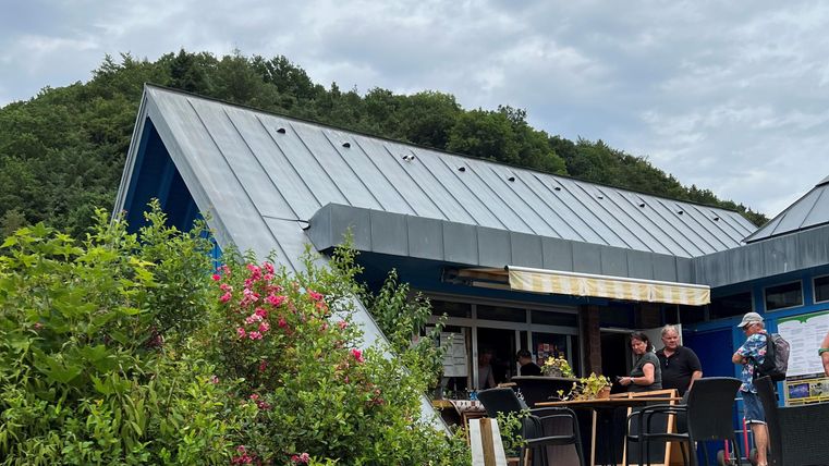 Ein modernes Gebäude mit einem schrägen Dach in einer natürlichen Umgebung. Menschen stehen draußen und genießen die Atmosphäre.