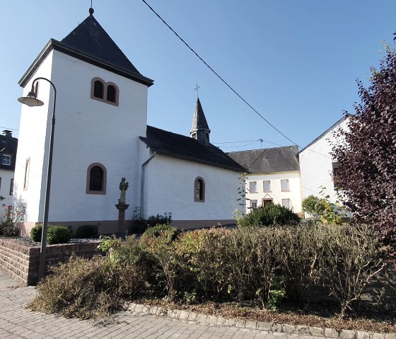 Die Kapelle St. Luzia steht in einem Dorf, umgeben von Häusern und Bäumen, unter einem klaren blauen Himmel., © TI Bitburger Land, Steffi Wagner
