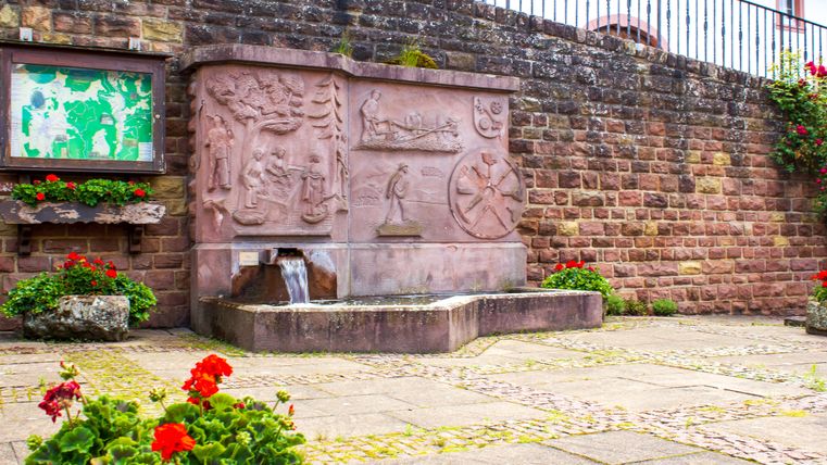 Een stenen dorpsfontein met reliëfs en stromend water, omringd door rode bloemen en een informatiebord op een stenen muur.