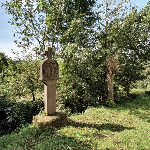 A stone cross stands on a green meadow, surrounded by trees and bushes. The sky is blue with a few clouds., © Tourist-Information Bitburger Land