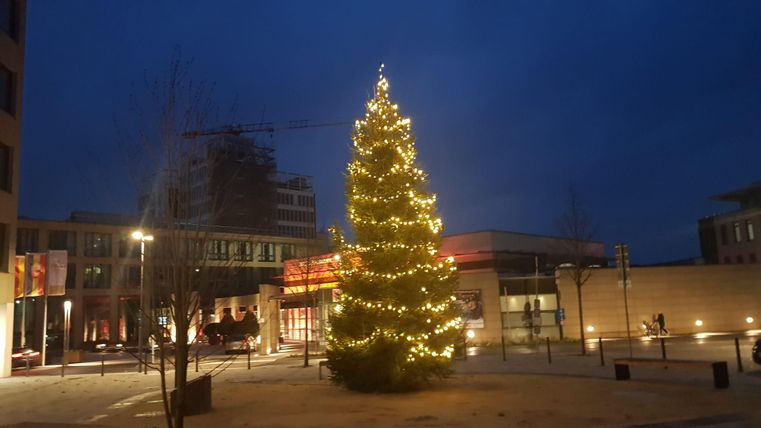 Ein festlich beleuchteter Weihnachtsbaum steht in einem urbanen Platz bei Dämmerung. Im Hintergrund sind moderne Gebäude und eine ruhige Atmosphäre zu sehen.