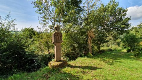 Croix de pierre sur une prairie, entourée d'arbres et d'arbustes, sous un ciel bleu.