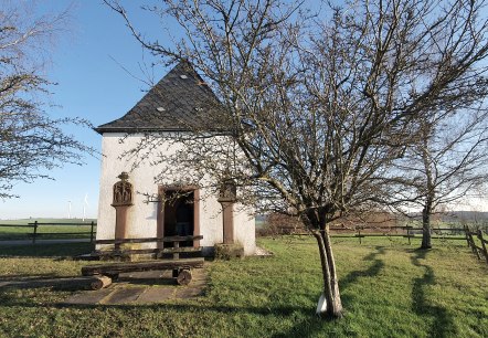 Eine kleine Kapelle auf einer grünen Wiese, umgeben von kahlen Bäumen. Im Hintergrund sind Windräder zu sehen., © TI Bitburger Land