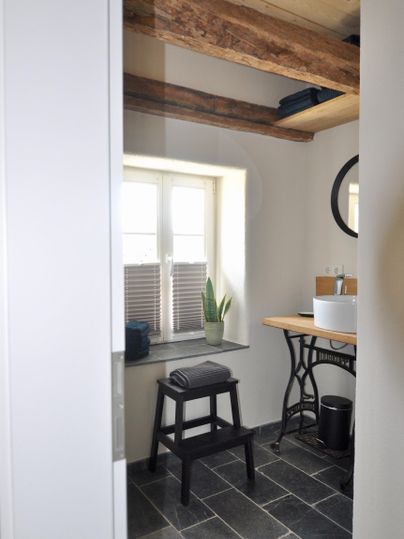 A modern bathroom with a window and a wooden chair. The walls are light, and the floors are tiled with dark tiles.