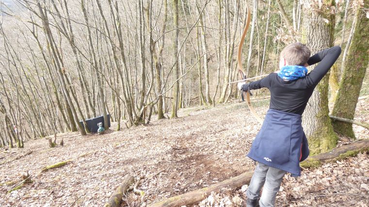 Ein Junge schießt mit einem Bogen in einem Wald. Die Umgebung ist von Bäumen und Laub geprägt.