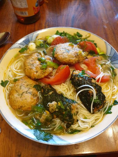 A bowl of noodle soup that contains meatballs, tomatoes, and fresh herbs. The ingredients are colorful and arranged appetizingly.