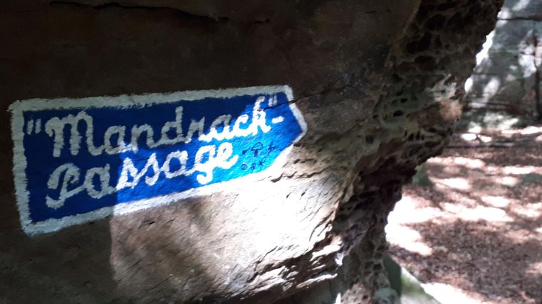 Ein blaue Schilder mit der Aufschrift "Mandrasch-Passage" zeigt den Weg. Es befindet sich an einer felsigen Oberfläche in einem Waldgebiet.