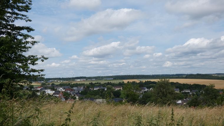 Landschaft mit Dorf, Feldern und Windrädern unter bewölktem Himmel.