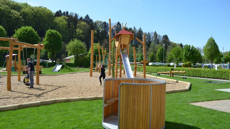 Ein schöner Kinderspielplatz mit einer Rutsche und verschiedenen Spielgeräten. Der Bereich ist von grünem Gras umgeben und es gibt viele Bäume im Hintergrund.