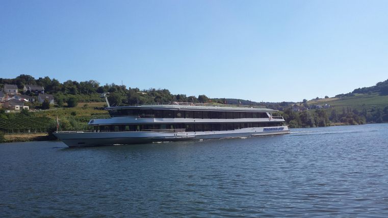 Ein elegantes Schiff fährt sanft auf einem ruhigen Gewässer. Im Hintergrund sind Hügel und grüne Landschaften sichtbar.