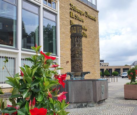 Ein Brunnen mit einer Skulptur vor einem modernen Gebäude. Lila und rote Blumen schmücken den Vordergrund.