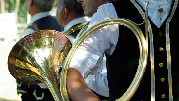 Een groep muzikanten draagt traditionele kleding en houdt instrumenten vast. Op de voorgrond is een groot blaasinstrument te zien.