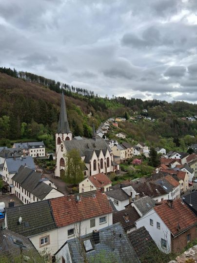 Eine malerische Dorfansicht mit einer Kirche im Vordergrund und sanften Hügeln im Hintergrund. Der Himmel ist bewölkt und die Häuser sind in verschiedenen Farben gehalten.