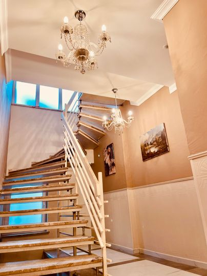 An elegant staircase with an open wooden staircase and two modern chandeliers. Artworks hang on the walls, while plenty of daylight comes through a large window.