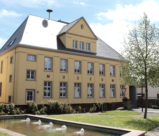 Yellow building of the Speicher local history museum with several windows. In the foreground a small fountain and a tree. The sky is blue with clouds., © TI Bitburger Land