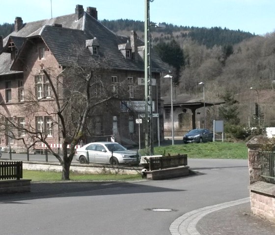 Historisch stationsgebouw in Zendscheid, omgeven door bomen en heuvels. Auto's parkeren voor het gebouw, dat gemaakt is van rode baksteen., © R. Schaal