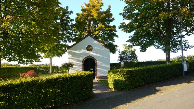 Eine kleine weiße Kapelle mit braunem Dach, umgeben von Bäumen und Hecken, bei sonnigem Wetter.