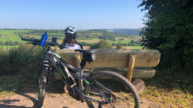 Person mit Helm sitzt auf einer Bank mit Blick auf eine grüne Landschaft, ein Fahrrad steht daneben.