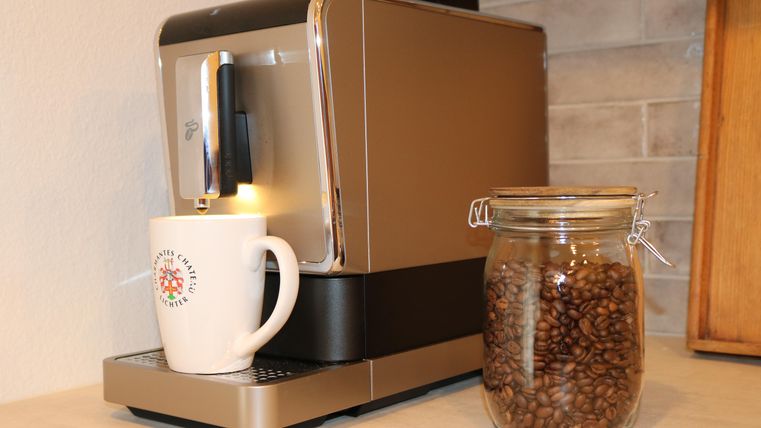 Een moderne koffiemachine staat op een werkblad naast een glas met koffiebonen. Een witte beker staat onder de uitloop van de machine.