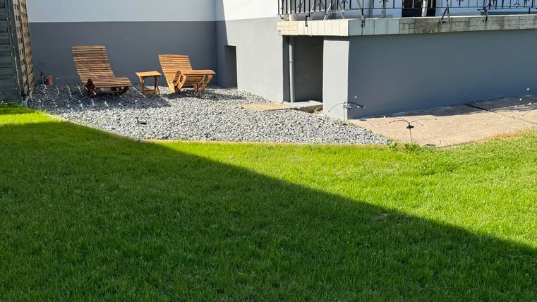 Une maison moderne avec un jardin bien entretenu. Deux chaises longues sont situées sur une zone de gravier à côté de la pelouse.