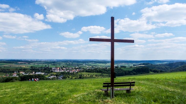 Ein großes Holzkreuz steht auf einer Wiese mit einer Bank davor. Im Hintergrund ist eine weite Landschaft mit einem Dorf zu sehen.