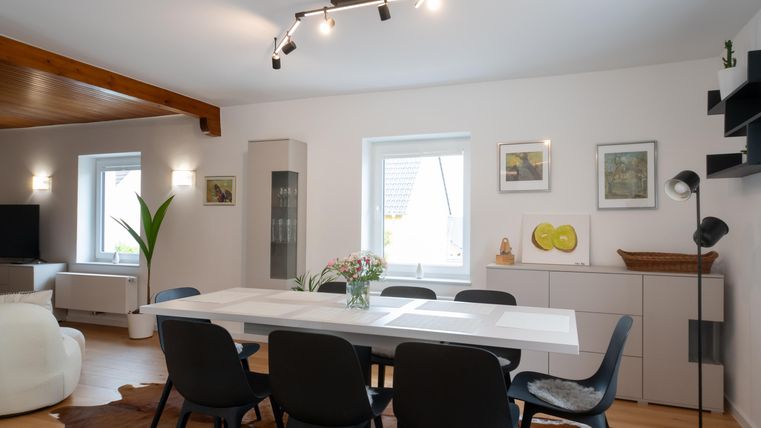 Een moderne eetkamer met een grote witte tafel en zwarte stoelen. De muren zijn licht geverfd en versierd met kunstwerken.