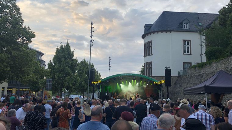 Een menigte bij een openluchtconcert voor een podium. Op de achtergrond zijn bomen en gebouwen zichtbaar, terwijl de lucht gedeeltelijk bewolkt is.