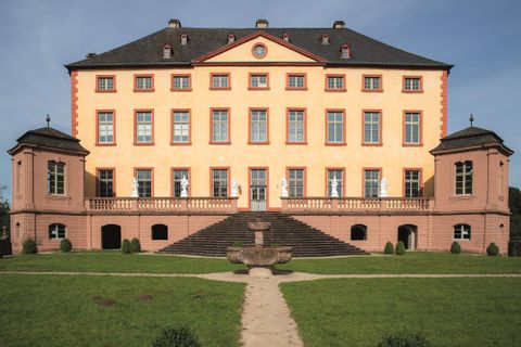 An impressive castle with an orange facade and large windows. It is surrounded by a well-maintained garden and offers a majestic view.