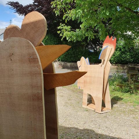 Two wooden animal sculptures are outdoors. One figure depicts a dog with a bright red tail, and the other figure is shaped like a chicken.