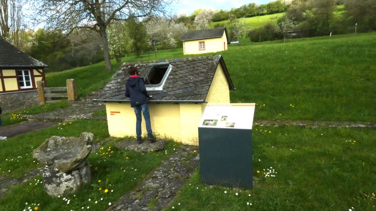 Ein Mann schaut in ein kleines gelbes Gebäude mit einem Schindeldach. Im Hintergrund sind weitere kleine Häuser auf einer grünen Wiese zu sehen.