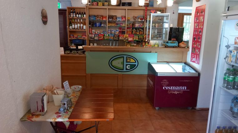 Ein gemütlicher Verkaufsraum mit Regalen voller Lebensmittel und Getränken. Im Vordergrund steht ein kleiner Tisch und eine Kühlschranktheke.