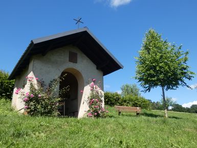 Kleine Kapelle im Grünen mit blühenden Rosen und Bank daneben.