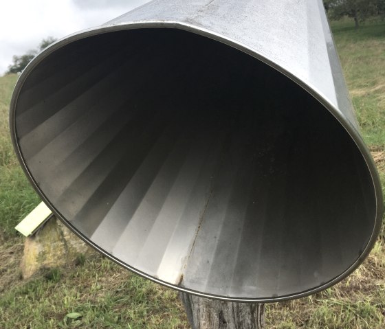 Un grand tube auditif en métal est posé sur une souche de bois dans une prairie verdoyante, prêt à amplifier les sons de l'environnement., © Tourist-Info Bitburger Land M.Mayer