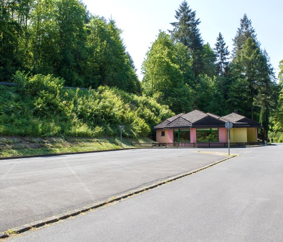 An empty parking lot with a small, colorful building in the background, surrounded by lush green trees., &copy; TI Bitburger Land