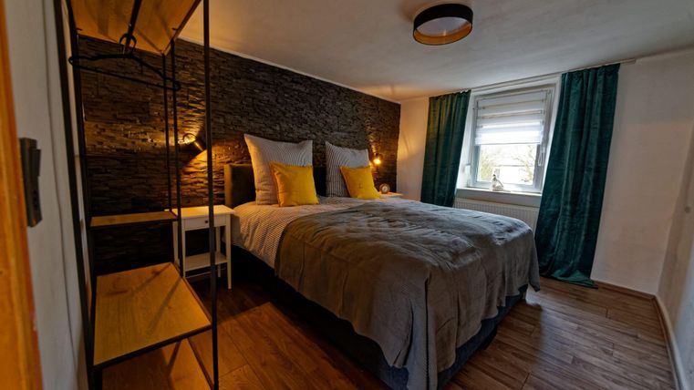 Ein gemütliches Schlafzimmer mit einem großen Bett, weichen Kissen und grünen Vorhängen. An der Wand befindet sich ein rustikales Design und ein heller Nachttisch.