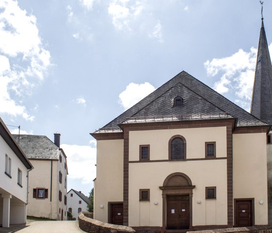 Die Pfarrkirche St. Peter in Neidenbach mit spitzem Turm, umgeben von Wohnhäusern, unter einem blauen Himmel mit Wolken., © TI Bitburger Land, M.Mayer